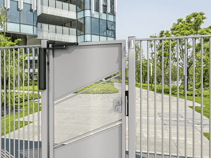 En moderne metalport med et geometrisk design og en Motoriseret og Automatiseret Portlukker til Fodgængerporte – VENUS er flankeret af et metalhegn. Porten er lidt åben og afslører en gangbro indeni. Træer og grønt græs er synlige i baggrunden sammen med en moderne glasbygning. Himlen er klar og solrig.