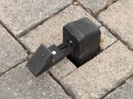 Et nærbillede af Universal portholder til porte/låger – RAL 9005 sidder mellem stenudlæggere arrangeret i et pænt mønster. Den sorte plastikclips er designet til at sikre genstande ved at gribe fat i de omgivende klodser og har en justerbar mekanisme til at stramme.