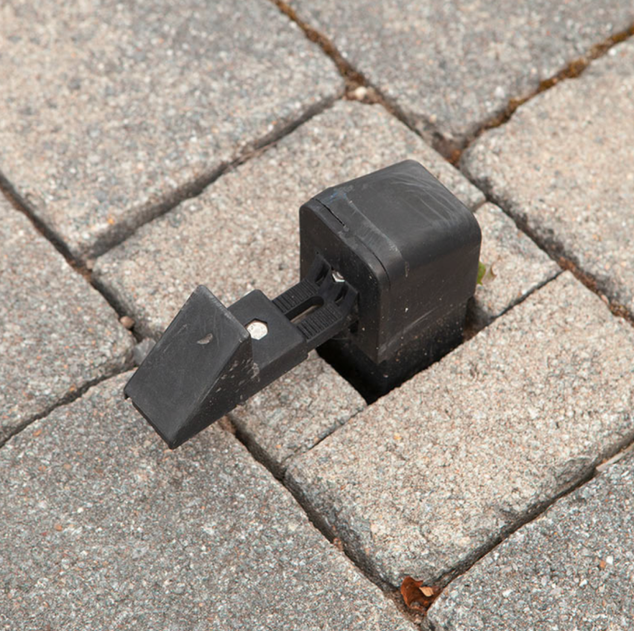 Et nærbillede af Universal portholder til porte/låger – RAL 9005 sidder mellem stenudlæggere arrangeret i et pænt mønster. Den sorte plastikclips er designet til at sikre genstande ved at gribe fat i de omgivende klodser og har en justerbar mekanisme til at stramme.