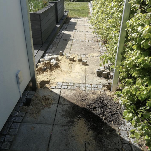 En smal sti foret med små, firkantede, grå belægningssten er delvist forstyrret. Nogle belægningssten er blevet fjernet, hvilket afslører sand og jord nedenunder. Til højre er der en frodig hæk langs stien, og der er en plantekasse synlig i baggrunden. En laage kan ses i nærheden.
