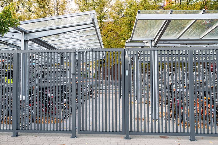 Den indhegnede cykelparkering har metalporte med VENUS motoriseret og automatiseret portlukker og gennemsigtigt tag. Talrige cykler er sikret inde, med efterårstræer i baggrunden. Fortovet udenfor er rent og velordnet.