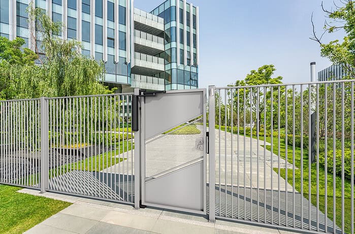 En moderne metalport med en VENUS Motoriseret og Automatiseret Portlukker til Fodgængerporte fører til en græsklædt, træbeklædt sti, der strækker sig til en moderne glaskontorbygning. Scenen er lys og solrig og afspejler en urban og grøn indstilling.