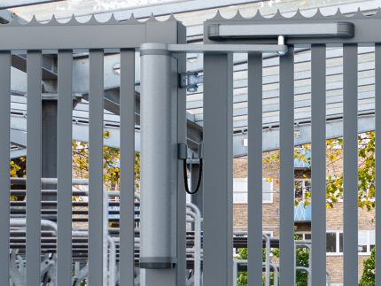 Nærbillede af VENUS motoriseret og automatiseret portlukker, der er sømløst fastgjort til en robust spalteport, med en overdækket gangbro, træer og bygning synlig gennem lamellerne, hvilket fremkalder et moderne bymiljø.