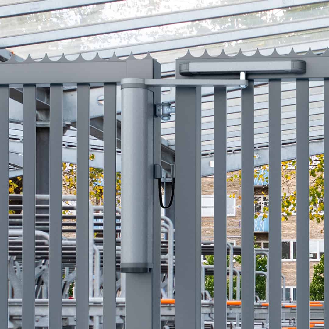 Nærbillede af VENUS motoriseret og automatiseret portlukker, der er sømløst fastgjort til en robust spalteport, med en overdækket gangbro, træer og bygning synlig gennem lamellerne, hvilket fremkalder et moderne bymiljø.