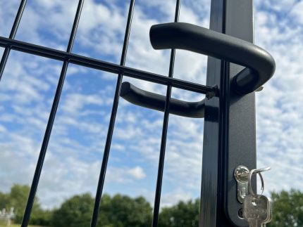 Nærbillede af et Håndtag sæt Sort Anodiseret Aluminium til Indsatslåse og en lås med nøgler hængende fra på et sort trådhegn. I baggrunden er der en græsmark med træer og en klar blå himmel fyldt med spredte hvide skyer.