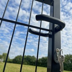 Nærbillede af et Håndtag sæt Sort Anodiseret Aluminium til Indsatslåse og en lås med nøgler hængende fra på et sort trådhegn. I baggrunden er der en græsmark med træer og en klar blå himmel fyldt med spredte hvide skyer.