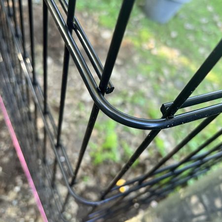 Nærbillede af hjørnet af et sort panelhegn, med en rød vandret tråd, der løber igennem det. Baggrunden er sløret og viser grønt græs og noget snavs. Sollys kaster skygger på 2D Panelhegn, og den overordnede scene giver en fornemmelse af udendørs omgivelser, der ligner billeder fundet i Montagevejledning-vejledninger.