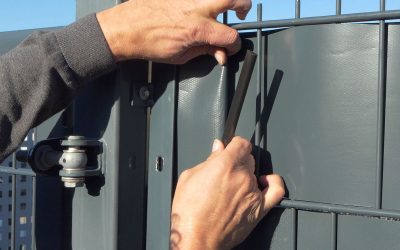 Nærbillede af en persons hænder, der installerer en strimmel sort gummi på kanten af et gråt metalpanel. Den ene hånd holder gummilisten åben, mens den anden hånd presser den på panelet. A Klemmeskinner Sort PVC til Afskærmningsruller 1 stk. er synlig til venstre for metalpanelet.