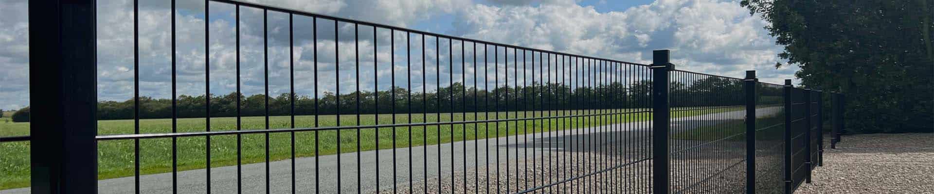 Et langt, sort metalhegn strækker sig ved siden af en grussti, der grænser op til et vidtstrakt grønt felt under en blå himmel med spredte skyer. Træer er synlige i den fjerne baggrund, og hegnet kaster skygge på stien.