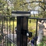 Nærbillede af en sort metalhegnspæl med bolte, der fastgør trådhegn, og en ZQUARE LED Stolpehat – Elegant Hegnsbelysning til Metalstolper, udendørs i en have med træer, grønne områder og en sløret statue i baggrunden.