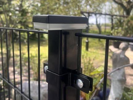Nærbillede af en sort metalhegnspæl med bolte, der fastgør trådhegn, og en ZQUARE LED Stolpehat – Elegant Hegnsbelysning til Metalstolper, udendørs i en have med træer, grønne områder og en sløret statue i baggrunden.