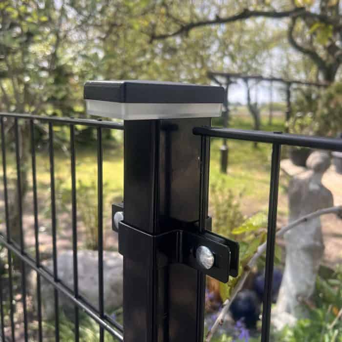 Nærbillede af en sort metalhegnspæl med bolte, der fastgør trådhegn, og en ZQUARE LED Stolpehat – Elegant Hegnsbelysning til Metalstolper, udendørs i en have med træer, grønne områder og en sløret statue i baggrunden.