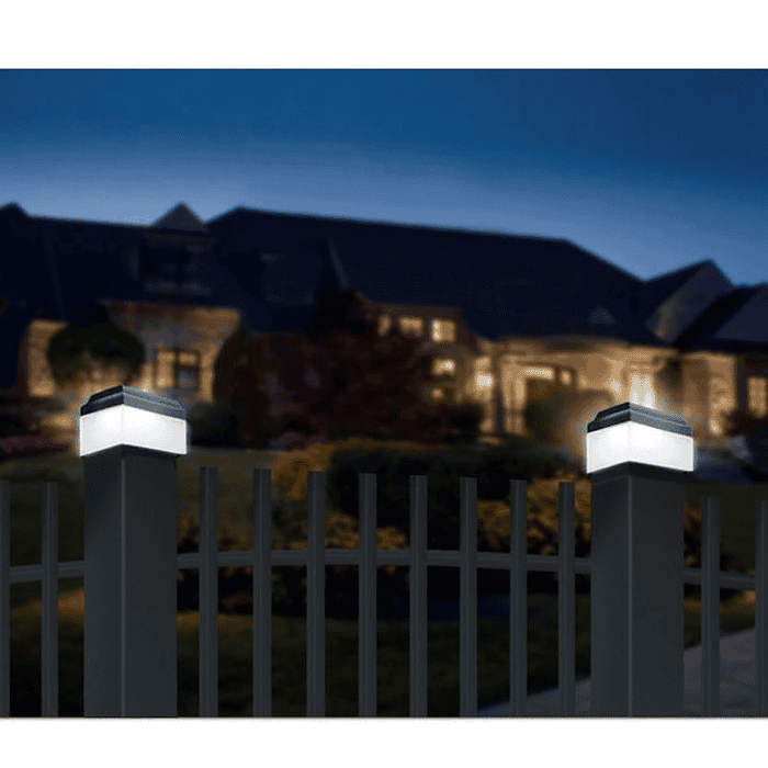 En natlig udsigt over et stort hus med varm indendørs belysning og en velholdt have. I forgrunden er der et metalhegn med to oplyste hegnsstolper med Solcelledrevet LED-lampe til Hegnsstolper, der tilføjer et blødt skær til scenen. Himlen er mørkeblå, hvilket indikerer aften eller nat.