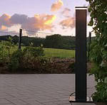 En sort lodret udendørs lysarmatur, såsom ZQUARE LED-Lys stolpehat til hegnsbelysning med metalstolper, står på en brolagt gangbro ved siden af en have med buske og vinstokke. I baggrunden er en græsmark og hegn oplyst under en himmel med orange skyer ved solnedgang.