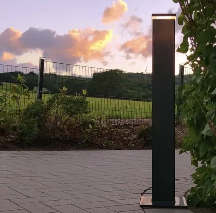 En sort lodret udendørs lysarmatur, såsom ZQUARE LED-Lys stolpehat til hegnsbelysning med metalstolper, står på en brolagt gangbro ved siden af en have med buske og vinstokke. I baggrunden er en græsmark og hegn oplyst under en himmel med orange skyer ved solnedgang.