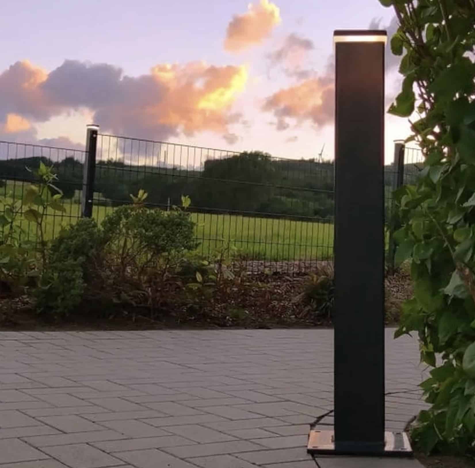 En sort lodret udendørs lysarmatur, såsom ZQUARE LED-Lys stolpehat til hegnsbelysning med metalstolper, står på en brolagt gangbro ved siden af en have med buske og vinstokke. I baggrunden er en græsmark og hegn oplyst under en himmel med orange skyer ved solnedgang.