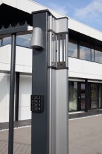 En moderne portstolpe med Design LED Belysning til Hegn og Porte - Locinox TRICONE numerisk tastatur indgangssystem og et LED-monteret lysarmatur står foran en moderne bygning med store vinduer og hvid facade. Tastaturet indeholder nummererede knapper 1-9, 0 og symbolerne CL og #.