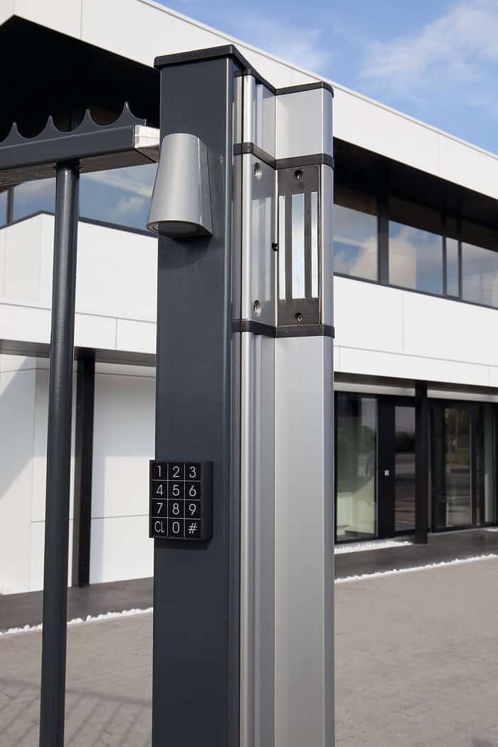 En moderne portstolpe med Design LED Belysning til Hegn og Porte - Locinox TRICONE numerisk tastatur indgangssystem og et LED-monteret lysarmatur står foran en moderne bygning med store vinduer og hvid facade. Tastaturet indeholder nummererede knapper 1-9, 0 og symbolerne CL og #.