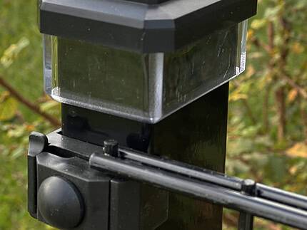 Et nærbillede af en Solcelledrevet LED-lampe fastgjort til hegnsstolper, med en gennemsigtig bundsektion. Placeret udendørs står den midt i levende grønt græs og delvist slørede planter og blander funktionalitet med naturen i et slankt 60x40 mm design.