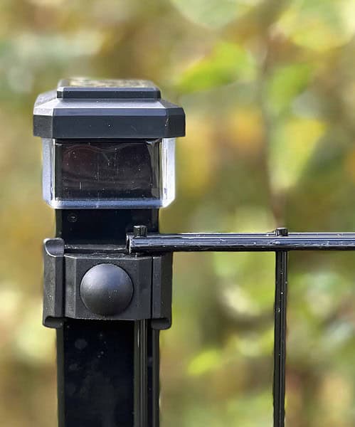 Nærbillede af en Solcelledrevet LED-lampe til Hegnsstolper i slank sort anbragt på en metalhegnspæl, blidt lysende mod en sløret baggrund af frodigt grønt løv.