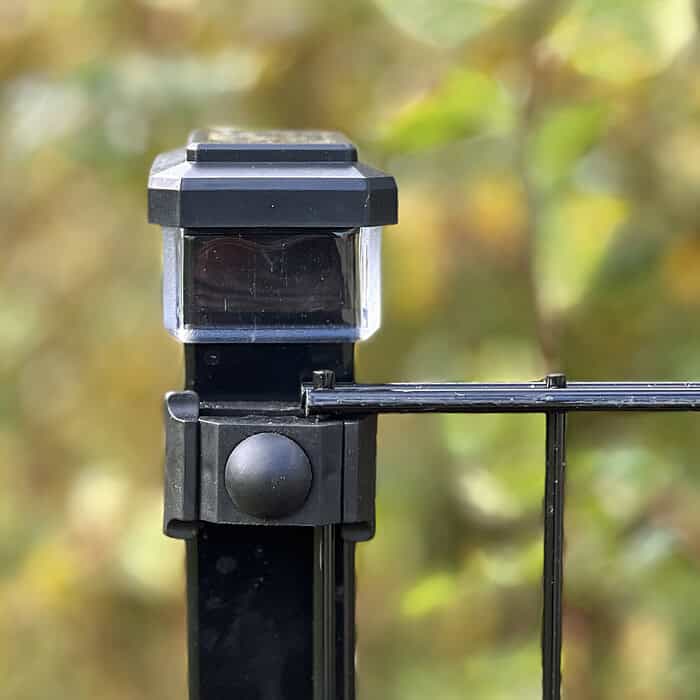 Nærbillede af en Solcelledrevet LED-lampe til Hegnsstolper i slank sort anbragt på en metalhegnspæl, blidt lysende mod en sløret baggrund af frodigt grønt løv.