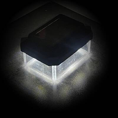 En lille rektangulær sort boks, der ligner en Solcelledrevet LED-lampe til Hegnsstolper, udsender et skarpt hvidt lys nedenunder, og kaster en oplyst refleksion på en mørk struktureret overflade. Den centralt placerede æske skaber en glorieeffekt omkring sin base.