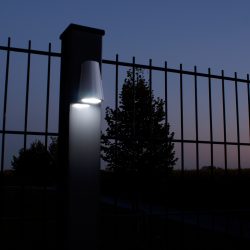 En tændt Design LED Belysning til Hegn og Porte - Locinox TRICONE er monteret på en søjle med lodrette metalstænger foran. Scenen, der foregår i skumringen eller tidlig aften, har en mørkere himmel og silhuet af træer i baggrunden. Området ser ud til at være en del af et hegn eller lågeindgang ved Hegn og Porte.