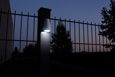 En tændt Design LED Belysning til Hegn og Porte - Locinox TRICONE er monteret på en søjle med lodrette metalstænger foran. Scenen, der foregår i skumringen eller tidlig aften, har en mørkere himmel og silhuet af træer i baggrunden. Området ser ud til at være en del af et hegn eller lågeindgang ved Hegn og Porte.