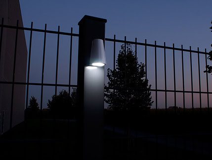 En tændt Design LED Belysning til Hegn og Porte - Locinox TRICONE er monteret på en søjle med lodrette metalstænger foran. Scenen, der foregår i skumringen eller tidlig aften, har en mørkere himmel og silhuet af træer i baggrunden. Området ser ud til at være en del af et hegn eller lågeindgang ved Hegn og Porte.