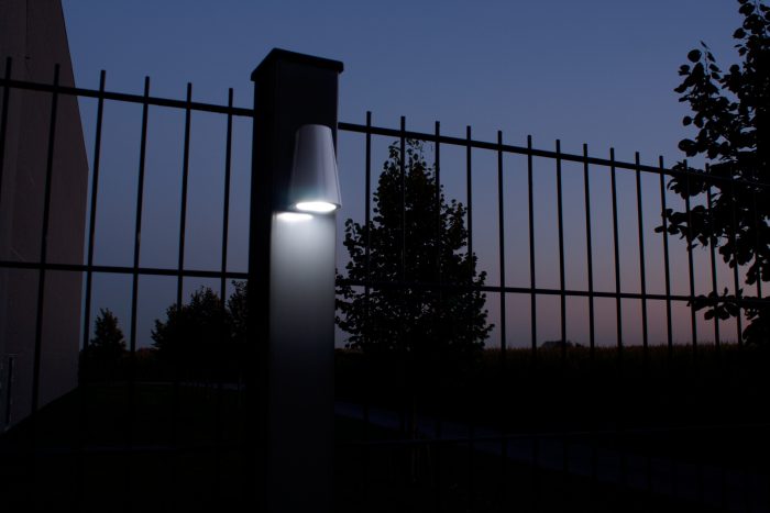En tændt Design LED Belysning til Hegn og Porte - Locinox TRICONE er monteret på en søjle med lodrette metalstænger foran. Scenen, der foregår i skumringen eller tidlig aften, har en mørkere himmel og silhuet af træer i baggrunden. Området ser ud til at være en del af et hegn eller lågeindgang ved Hegn og Porte.