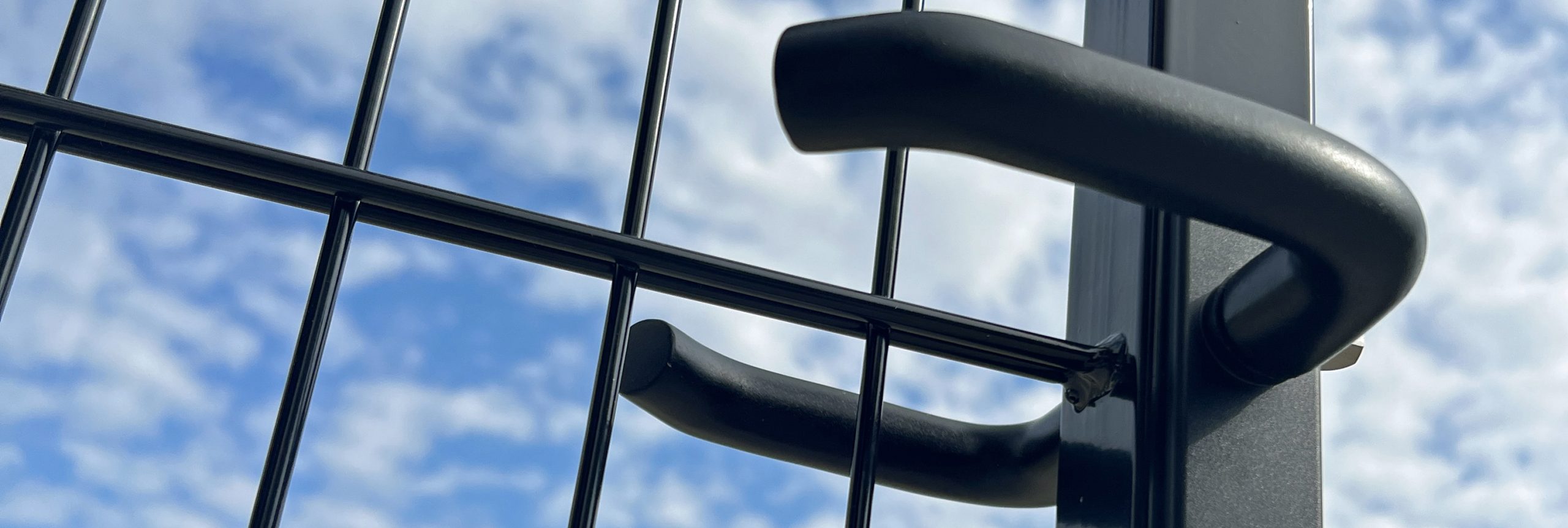 Nærbillede af to sorte metalhåndtag på en trådhegnslåge, på baggrund af en delvist overskyet blå himmel.