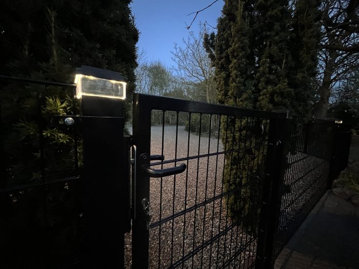 En sort metalport med lås har et stolpehat med lys – en solcelledrevet LED-lampe til hegnsstolper – der lyser sagte, idet det åbner sig ud til en grussti omkranset af træer og buske under den svage, tidlige aftenhimmel.
