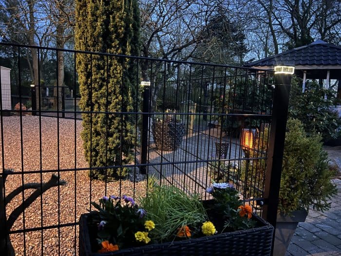 En have i skumringen med et metalhegn, blomsterkasse, grussti, høje træer og et lysthus. Stolpehat med lys - Solcelledrevet LED-lampe til Hegnsstolper oplyser blødt stien og hegnet for en hyggelig atmosfære.