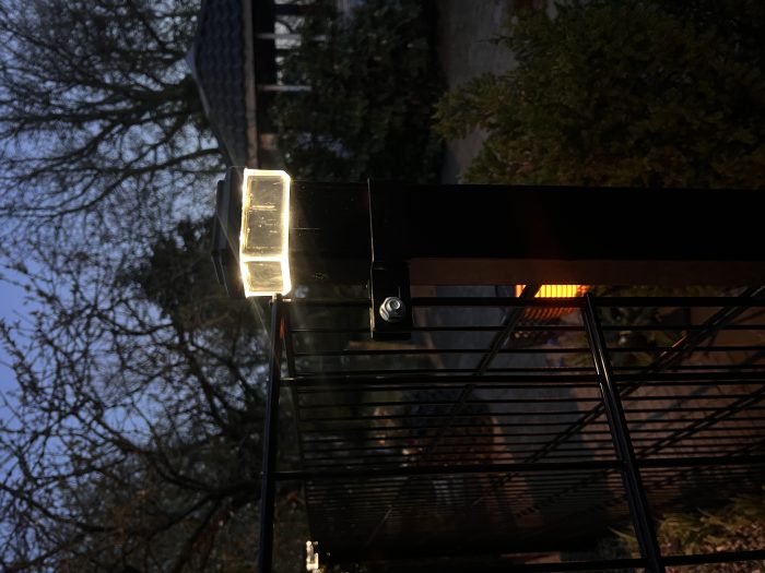 Et nærbillede af en Stolpehat med lys, en solcelledrevet LED-lampe til hegnsstolper, monteret på toppen af et sort metalhegn ved skumring. Træer og et lysthus anes i baggrunden.
