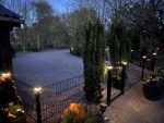 En blødt oplyst have i skumringen med en grusindkørsel, høje stedsegrønne træer og et sort metalhegn med Stolpehat med lys—solcelledrevet LED-lampe til hegnsstolper. Potteplanter omkranser murstensterrassen; bladløse træer står i baggrunden.