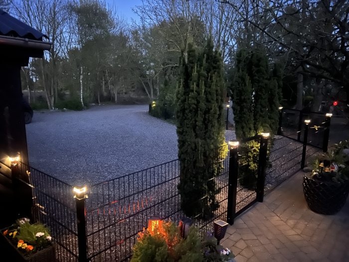 En blødt oplyst have i skumringen med en grusindkørsel, høje stedsegrønne træer og et sort metalhegn med Stolpehat med lys—solcelledrevet LED-lampe til hegnsstolper. Potteplanter omkranser murstensterrassen; bladløse træer står i baggrunden.