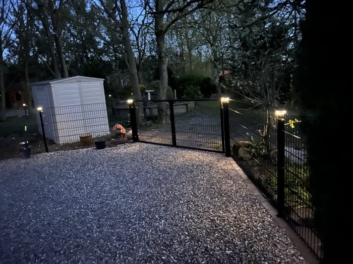 Ved skumringstid har en indhegnet have Stolpehat med lys - Solcelledrevet LED-lampe til Hegnsstolper på portpælene, en grusindkørsel foran, et hvidt skur til venstre og træer i baggrunden.