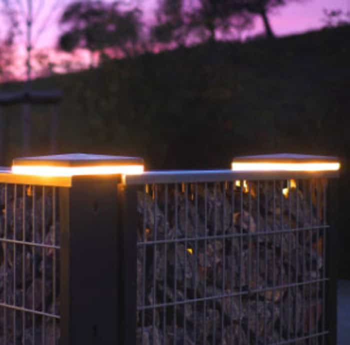 Et hegn bygget af metalriste fyldt med sten er blødt oplyst af to ZQUARE LED-Lys stolpehat til hegnsbelysning med metalstolper, der kaster et blidt skær i skumringen. Det slørede landskab i baggrunden er sat mod en lilla himmel.