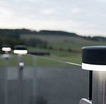 En fredfyldt udendørs scene med ZQUARE LED-Lys stolpehat til hegnsbelysning med metalstolper, minimalistiske sorte og sølvfarvede lys monteret på et rækværk, med udsigt over et sløret landskab af marker og træer under en overskyet himmel. Fokus er på lysene, hvilket skaber en slank og moderne stemning.