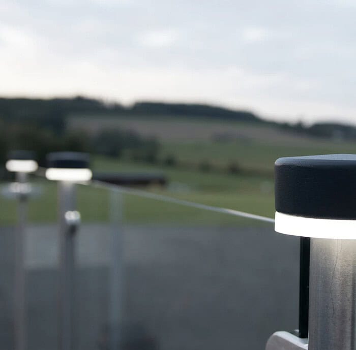 En fredfyldt udendørs scene med ZQUARE LED-Lys stolpehat til hegnsbelysning med metalstolper, minimalistiske sorte og sølvfarvede lys monteret på et rækværk, med udsigt over et sløret landskab af marker og træer under en overskyet himmel. Fokus er på lysene, hvilket skaber en slank og moderne stemning.