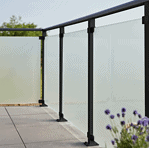 En moderne udendørs altan med 90x91x6mm matte hærdet glaspaneler og sort metal gelænder. Altangulvet er flisebelagt, og i forgrunden ses lilla blomster, der tilføjer et strejf af natur til scenen. Baggrunden viser en klar, solrig himmel med sorte beslag, der subtilt fremhæver det slanke design.