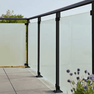 En moderne udendørs altan med 90x91x6mm matte hærdet glaspaneler og sort metal gelænder. Altangulvet er flisebelagt, og i forgrunden ses lilla blomster, der tilføjer et strejf af natur til scenen. Baggrunden viser en klar, solrig himmel med sorte beslag, der subtilt fremhæver det slanke design.