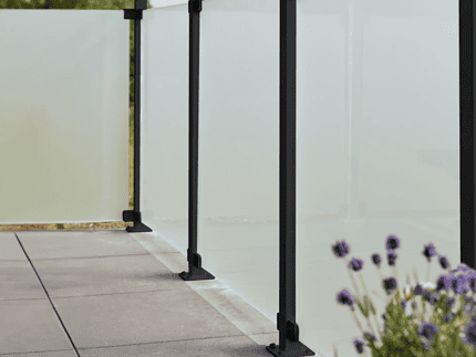 En moderne udendørs altan med 90x91x6mm matte hærdet glaspaneler og sort metal gelænder. Altangulvet er flisebelagt, og i forgrunden ses lilla blomster, der tilføjer et strejf af natur til scenen. Baggrunden viser en klar, solrig himmel med sorte beslag, der subtilt fremhæver det slanke design.