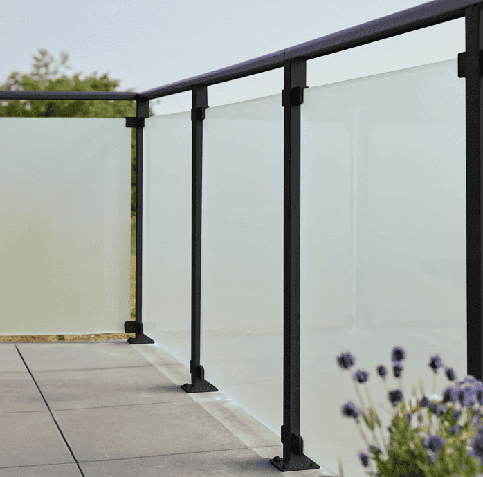 En moderne udendørs altan med 90x91x6mm matte hærdet glaspaneler og sort metal gelænder. Altangulvet er flisebelagt, og i forgrunden ses lilla blomster, der tilføjer et strejf af natur til scenen. Baggrunden viser en klar, solrig himmel med sorte beslag, der subtilt fremhæver det slanke design.