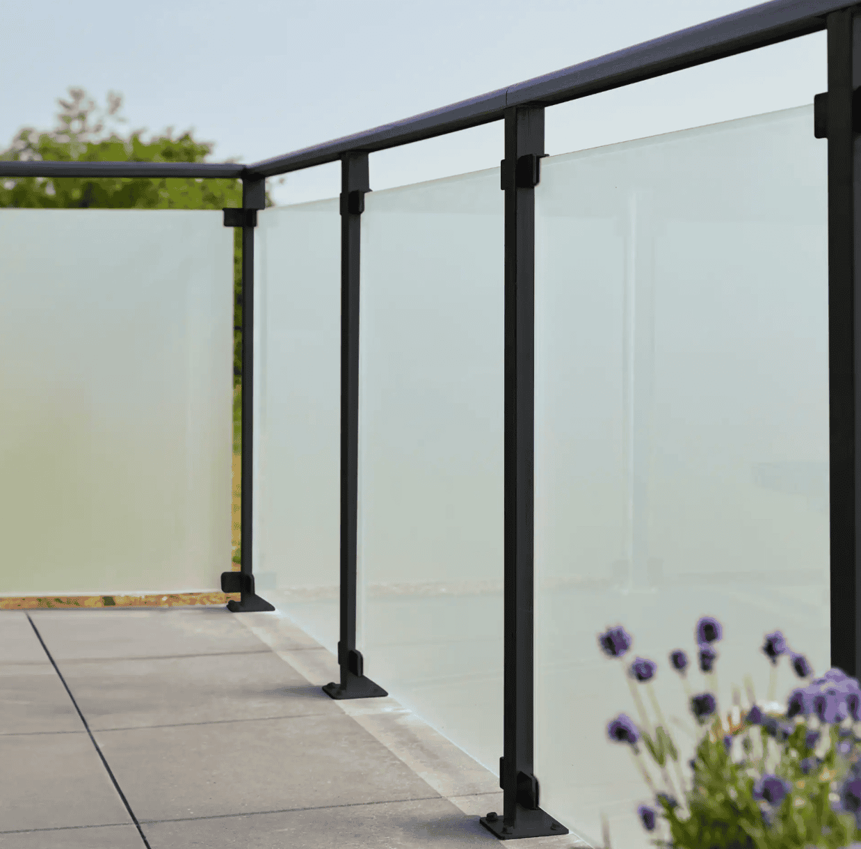 En moderne udendørs altan med 90x91x6mm matte hærdet glaspaneler og sort metal gelænder. Altangulvet er flisebelagt, og i forgrunden ses lilla blomster, der tilføjer et strejf af natur til scenen. Baggrunden viser en klar, solrig himmel med sorte beslag, der subtilt fremhæver det slanke design.