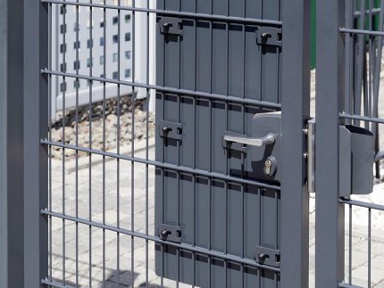 Et nærbillede af en metalsikkerhedslåge med vandrette og lodrette stænger med "TREND" lågeplade til indgrebssikring. Porten er sikret med hængelås og lås. I baggrunden er der noget hegn og grus på jorden, hvilket indikerer udendørs omgivelser.