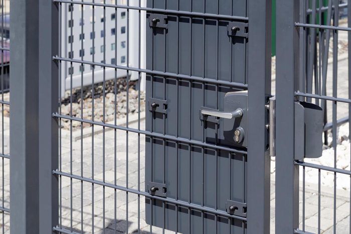 Et nærbillede af en metalsikkerhedslåge med vandrette og lodrette stænger med "TREND" lågeplade til indgrebssikring. Porten er sikret med hængelås og lås. I baggrunden er der noget hegn og grus på jorden, hvilket indikerer udendørs omgivelser.