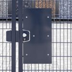 Et nærbillede af den sorte Lågeplade til Indgrebssikring metal "TREND" med et mesh-design, der fremhæver dens robuste sikkerhedsfunktioner. Porten inkluderer en rektangulær sort plade på den ene side og et sølv dørhåndtag med nøglehul. Bag porten ses stenkonstruktioner og en brolagt jordoverflade.