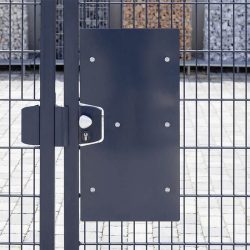 Et nærbillede af den antracit Lågeplade til Indgrebssikring metal "TREND" med et mesh-design, der fremhæver dens robuste sikkerhedsfunktioner. Porten inkluderer en rektangulær sort plade på den ene side og et sølv dørhåndtag med nøglehul. Bag porten ses stenkonstruktioner og en brolagt jordoverflade.