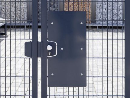 Et nærbillede af den antracit Lågeplade til Indgrebssikring metal "TREND" med et mesh-design, der fremhæver dens robuste sikkerhedsfunktioner. Porten inkluderer en rektangulær sort plade på den ene side og et sølv dørhåndtag med nøglehul. Bag porten ses stenkonstruktioner og en brolagt jordoverflade.
